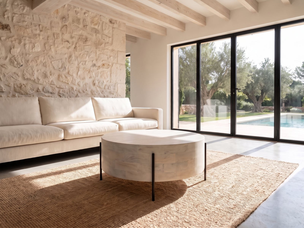 Lucignano Whitewashed Mango Drum Coffee Table seen from the side, on a jute rug in front of a linen sofa in a poolside villa