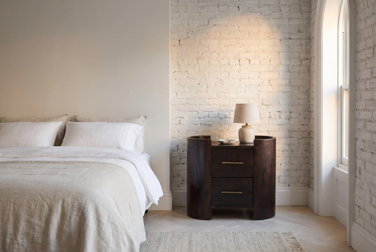 Macomer mocha mango oval nightstand with a ceramic lamp against a whitewashed brick wall in a Brooklyn bedroom with an arched window and a herringbone floor