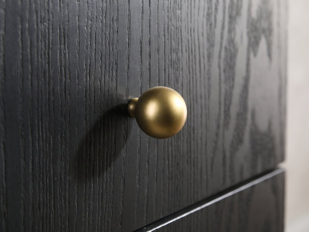 Close-up of the Malcesine 63-Inch Wire-Brushed Black 8-Drawer Dresser: a round soft-gold ball knob on the black wire-brushed grain of a drawer front