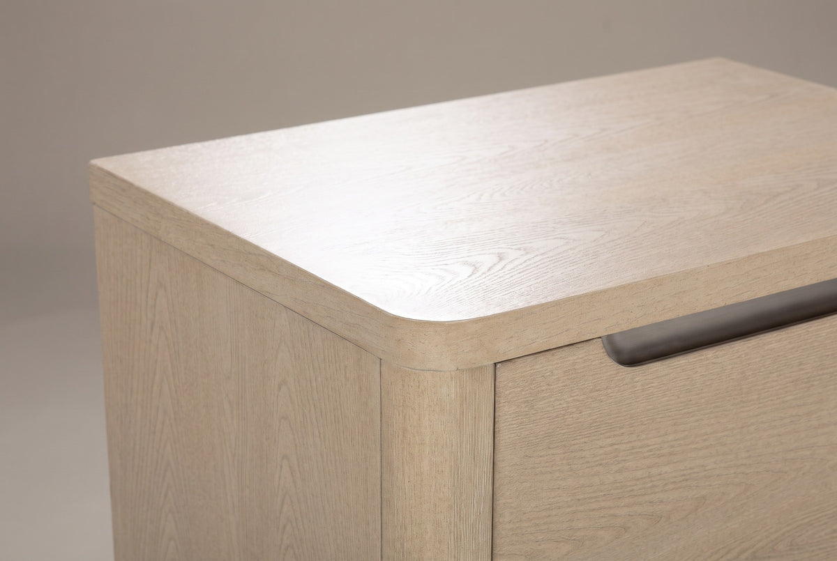 The top corner of the Mandas nightstand: a thick light elm top with a rounded front corner, the rounded corner post and the end of a nickel-lined scoop on the upper drawer