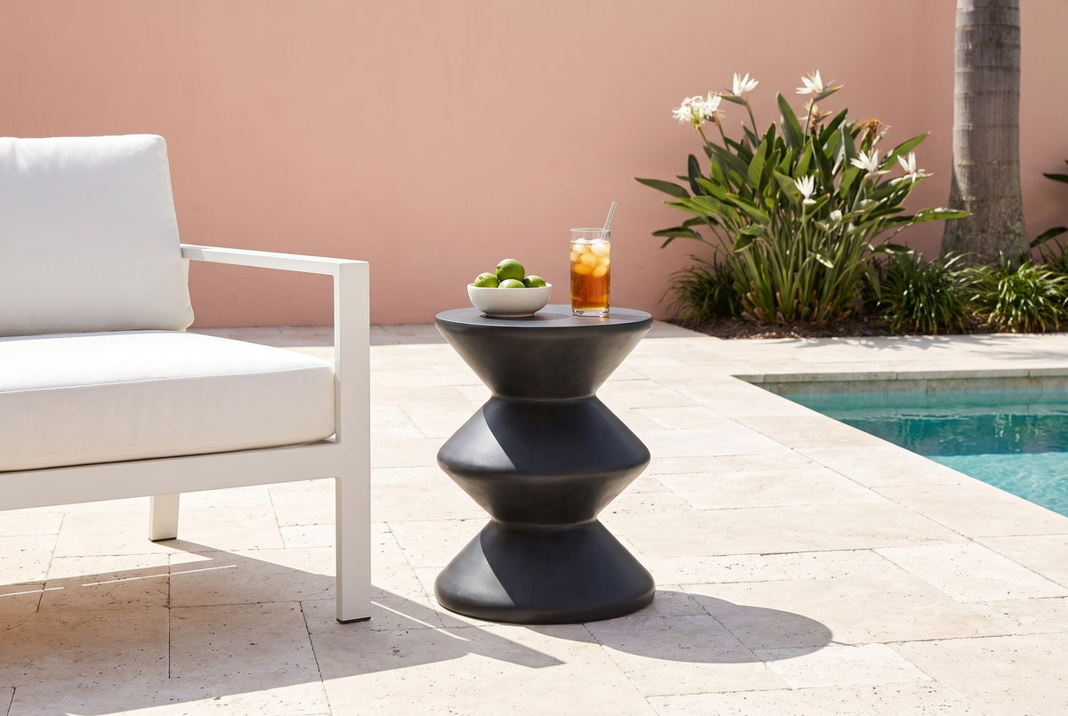 Marineo black concrete zigzag side table with a bowl of limes and a glass of iced tea beside a white lounge armchair on a travertine pool terrace in Naples, Florida, a coral-pink stucco wall and bird-of-paradise behind (chair and decor not included)