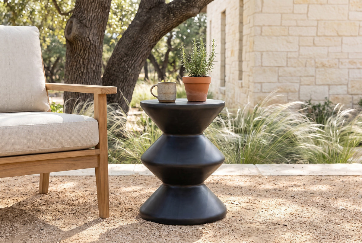 Marineo black concrete zigzag side table with a stoneware mug and a pot of rosemary beside a teak lounge chair on a decomposed granite patio in the Texas Hill Country, a cream limestone wall, feather grass and live oaks behind (chair and decor not included)