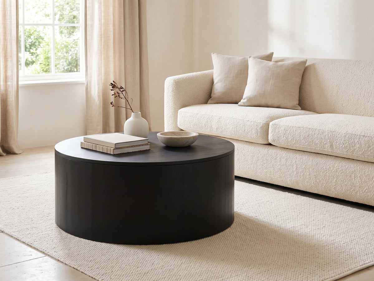 Marostica Black Mango Drum Coffee Table styled with books, a vase and a stone bowl in front of a cream sofa in a bright plaster room