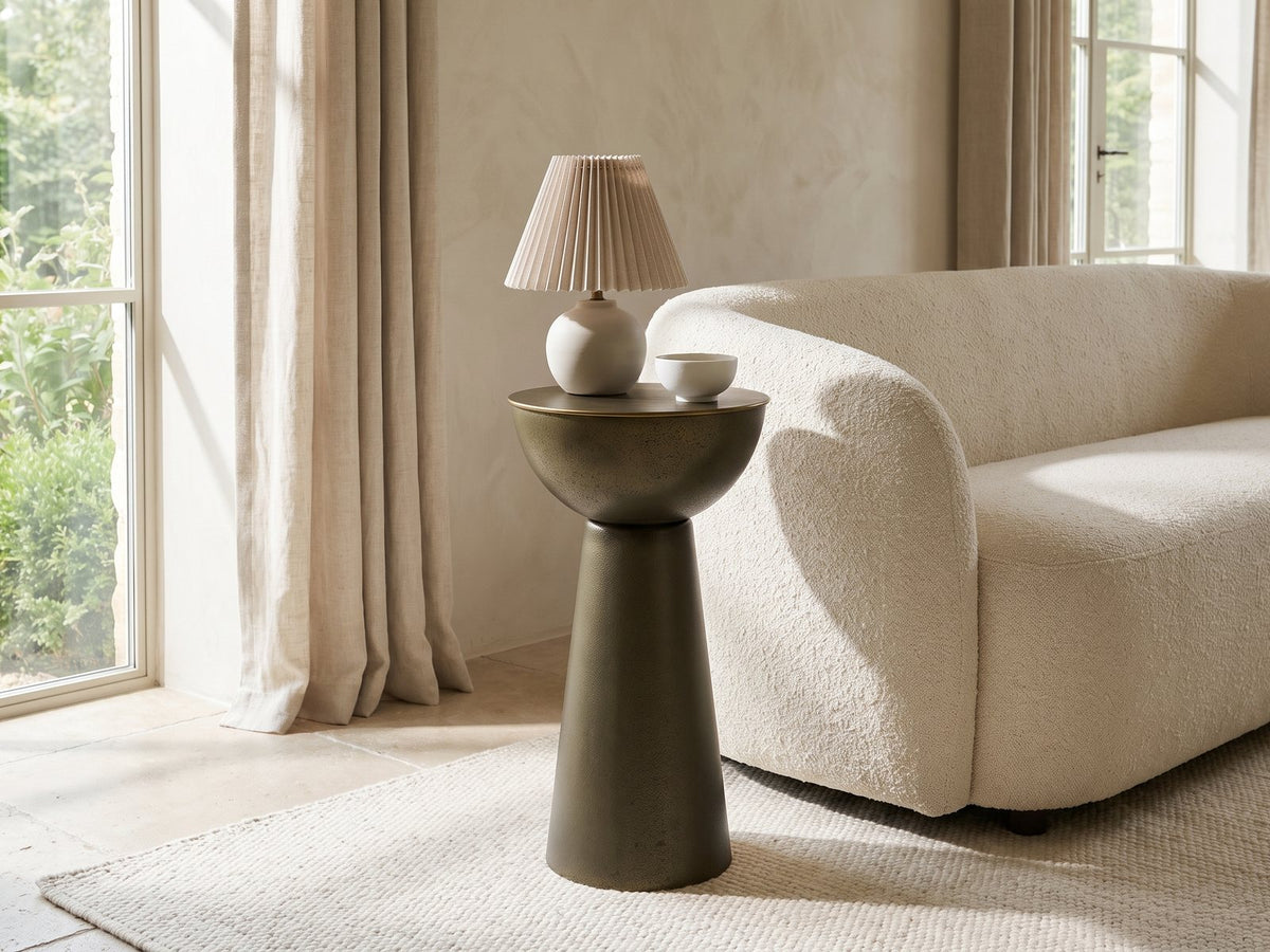 Melpignano Antique Gold Iron Pedestal Side Table with a lamp and a ceramic bowl beside a cream boucle sofa in a bright plaster room