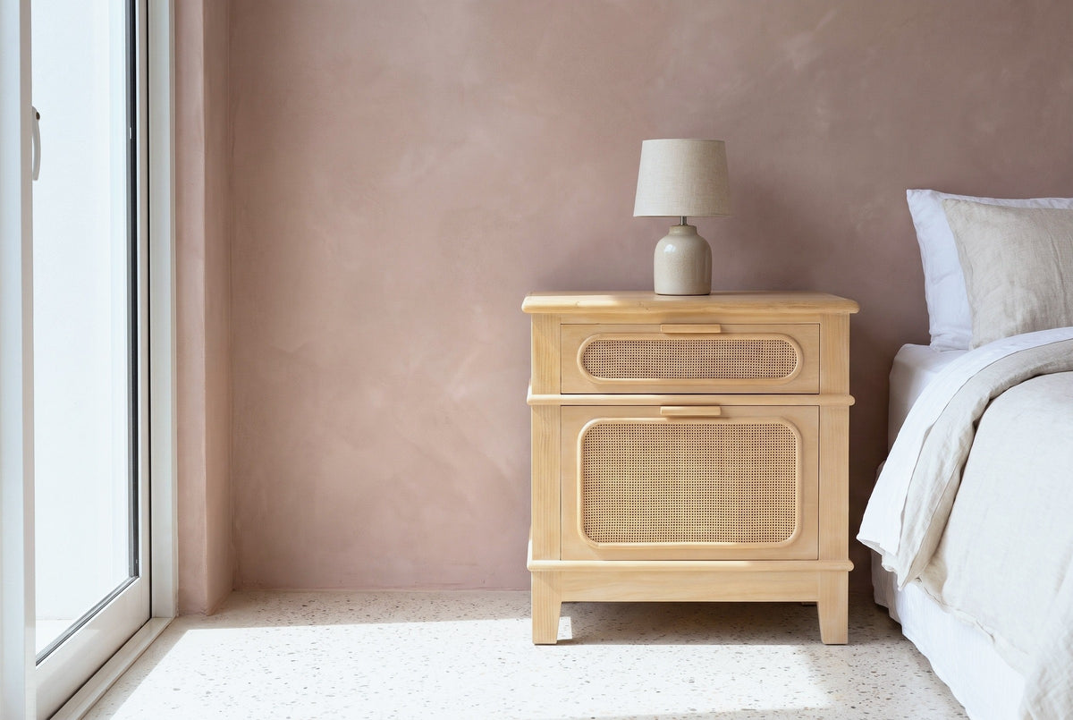 Mogoro pine and cane nightstand with a ceramic lamp beside a bed in a Los Angeles bedroom with a dusty-rose plaster wall, a terrazzo floor and a tall glass door
