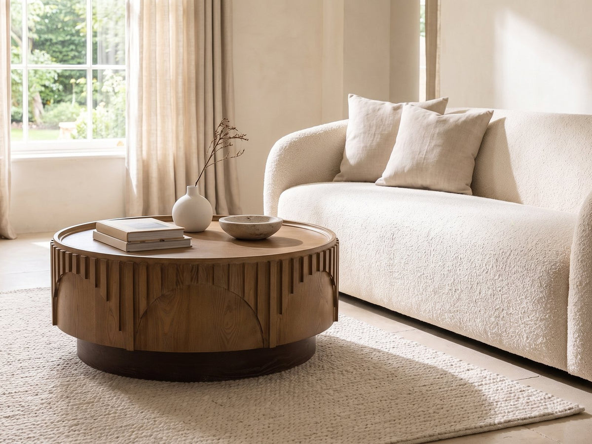 Mondovi 36-Inch Round Carved Limed Ash Drum Coffee Table seen from the side, styled with books, a vase and a stone bowl in front of a cream sofa in a bright plaster room
