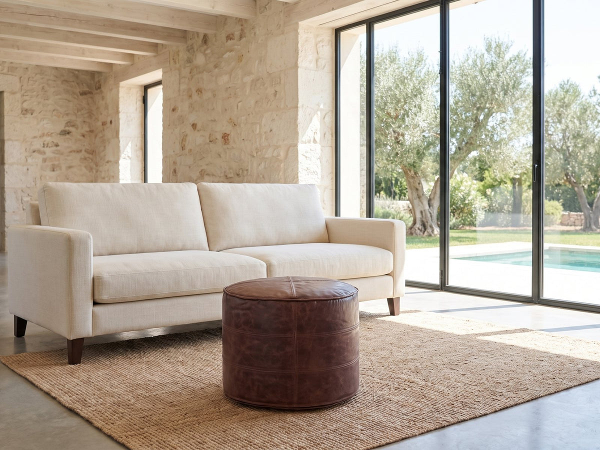 Moneglia Distressed Brown Top-Grain Buffalo Leather Round Pouf on a jute rug in front of a linen sofa in a poolside villa
