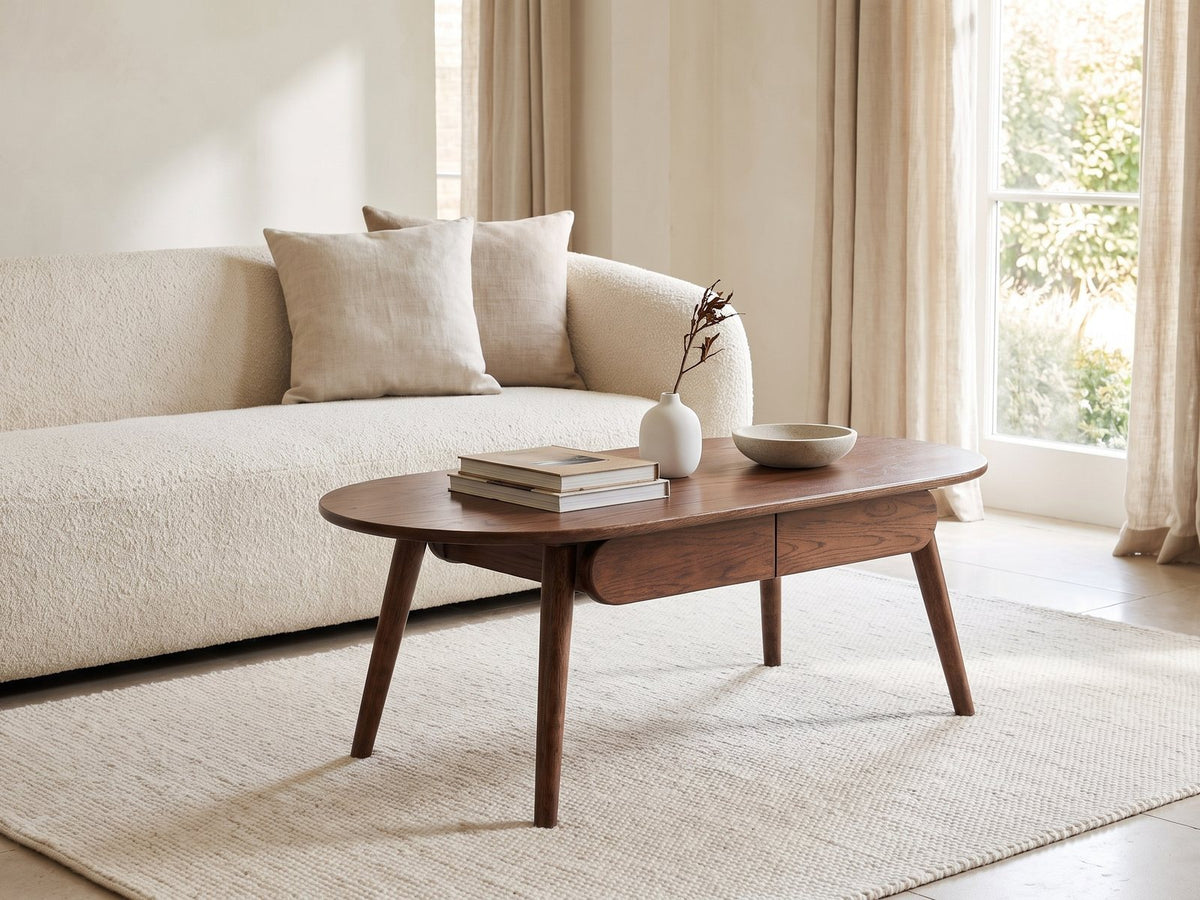 Neive 47-Inch Oval Walnut-Stained Solid Oak Coffee Table with 2 Drawers styled with books, a vase and a stone bowl in front of a cream sofa in a bright plaster room