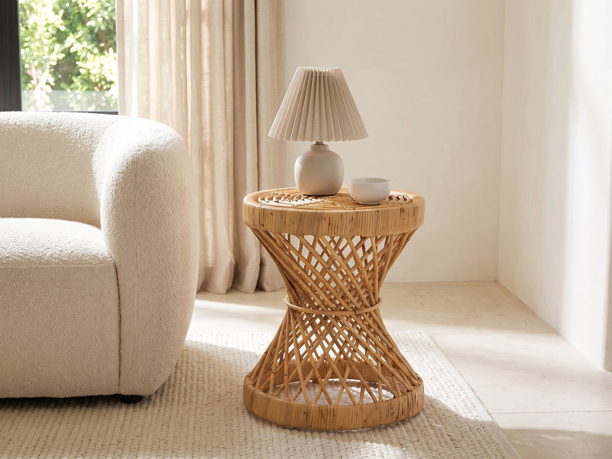 Neviano Natural Rattan Hourglass Side Table with a lamp and a ceramic bowl beside a cream boucle armchair in a bright plaster room