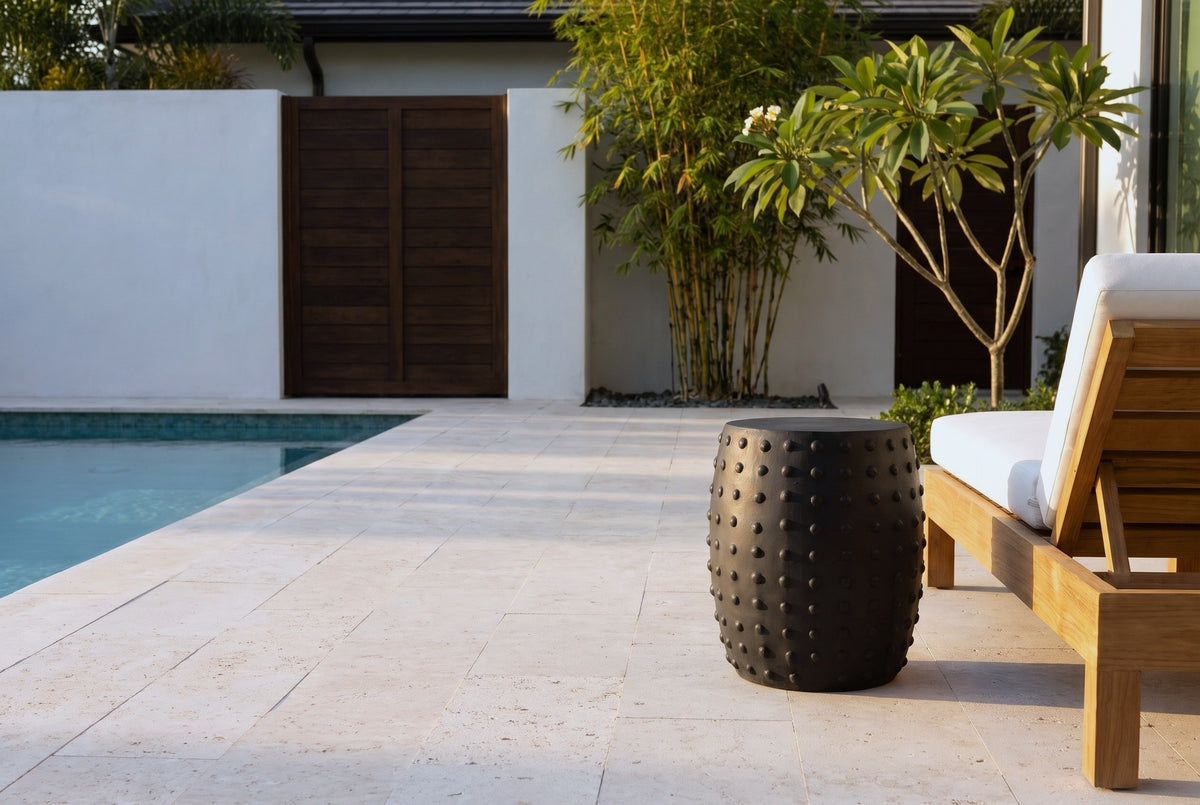 Nicolosi matte black studded concrete side table on a travertine pool deck in Naples, Florida, a white wall, bamboo and a teak lounger behind (lounger not included)