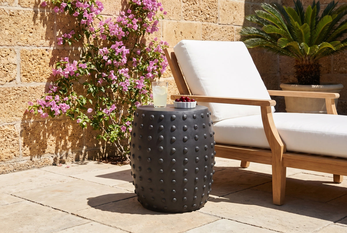 Nicolosi matte black studded side table in a St. Augustine courtyard with a coquina stone wall and bougainvillea, beside a teak lounger (lounger, glass and cherries not included)