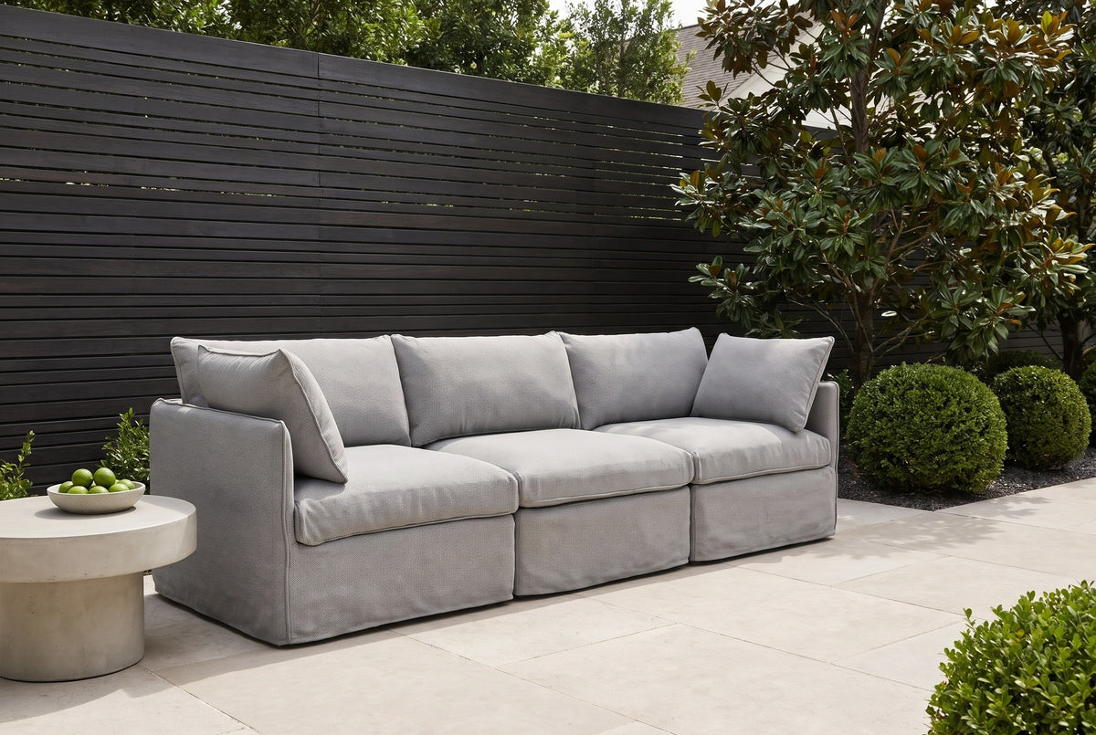 Nicosia grey slipcovered sofa on pale limestone pavers in a Houston garden, a charcoal slatted fence, boxwood and a magnolia behind (side table not included)