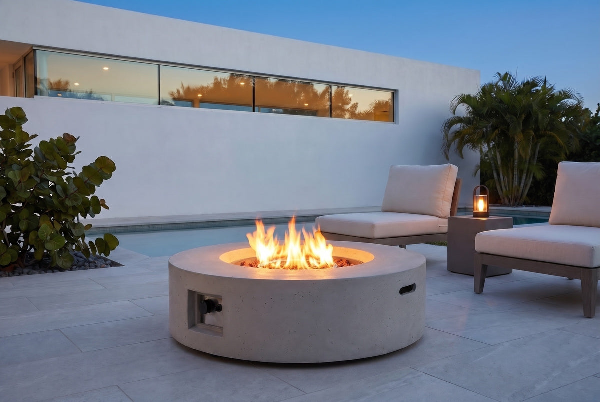 Noto fire pit in light gray concrete, lit on a pool terrace in Sarasota, Florida at blue hour, a white stucco wall with a band of glass and palms behind (chairs, side table and lantern not included)