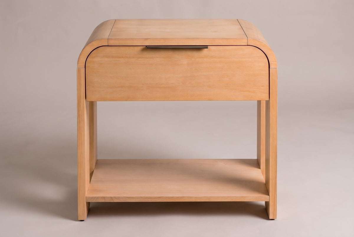The Nuoro Caramel Waterfall nightstand, front view on a warm grey studio backdrop: a waterfall top, one drawer with a slim bronze pull, four slab legs and an open shelf