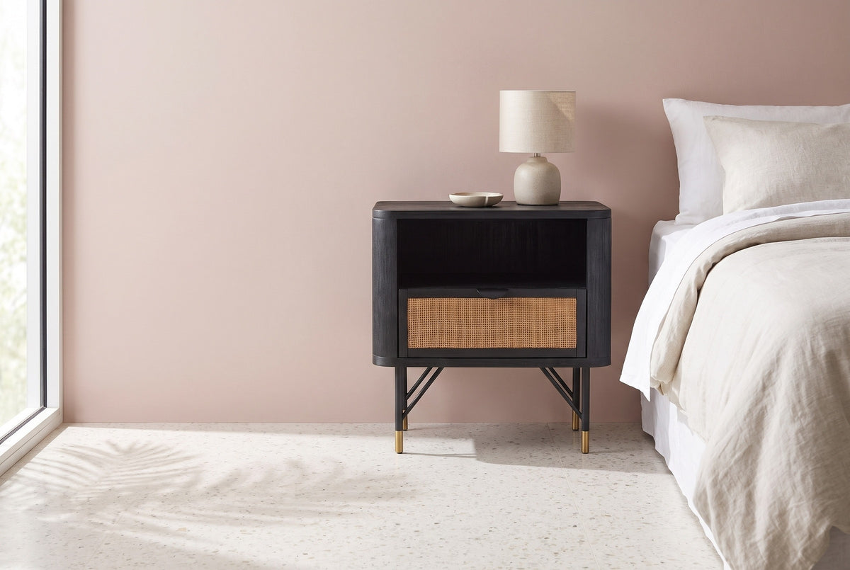 Olbia black acacia and rattan nightstand with a ceramic lamp beside a bed in a Palm Springs bedroom with a dusty blush pink plaster wall and a terrazzo floor