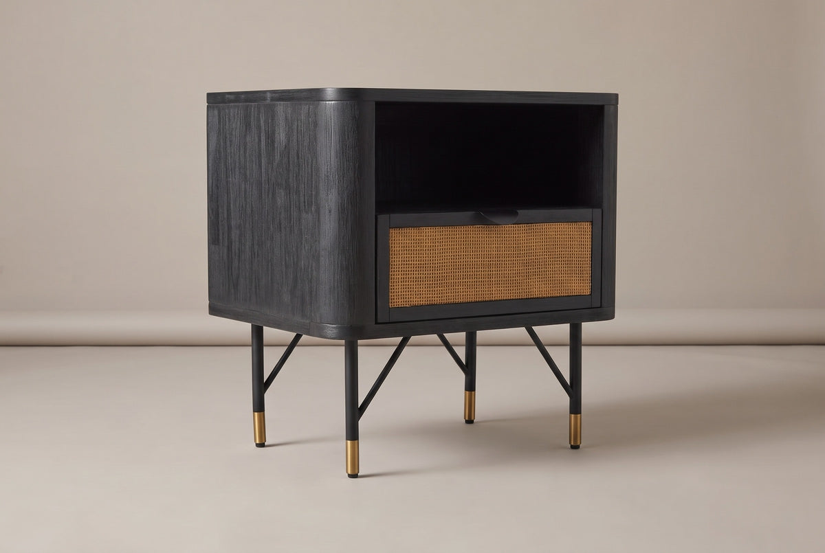The Olbia one-drawer nightstand in black acacia with an open niche, a rattan cane drawer front and four slim black legs with brass caps, three-quarter view on a light studio backdrop
