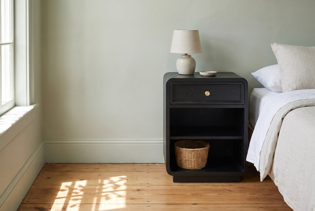 Oliena black rounded nightstand with a ceramic lamp and a woven basket beside a bed in a Charleston bedroom with a pale sage green plaster wall and a heart-pine floor
