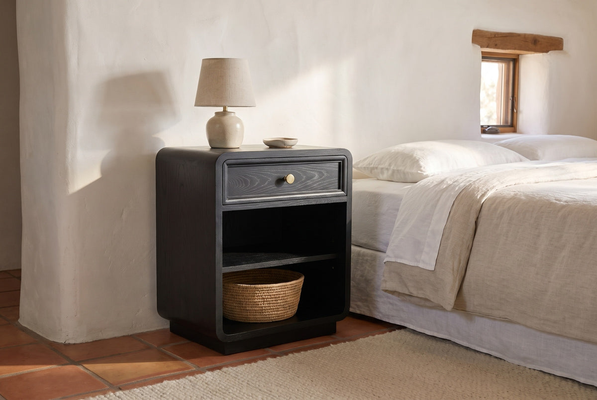 Oliena black rounded nightstand with a ceramic lamp and a woven basket against a white-washed adobe wall in a Santa Fe bedroom with a terracotta tile floor