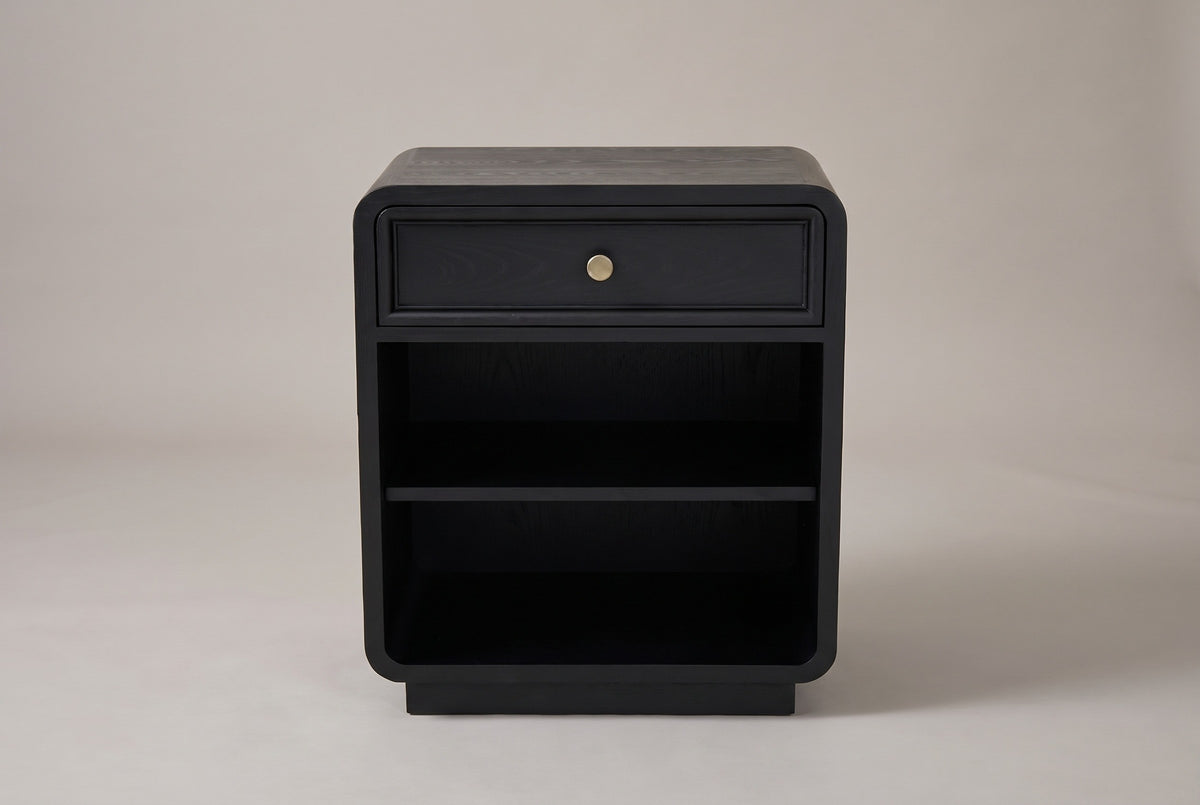 The Oliena black rounded nightstand, front view on a warm grey studio backdrop: one framed drawer with a brass knob, an open compartment with one shelf and a low plinth