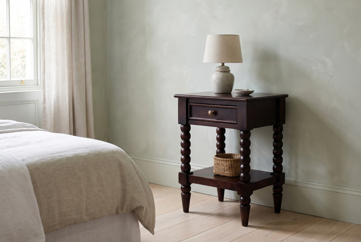 Orani dark winewood bobbin-leg nightstand with a ceramic lamp and a seagrass basket on the shelf beside a bed in a Charleston bedroom with a pale sage plaster wall and a pale oak floor