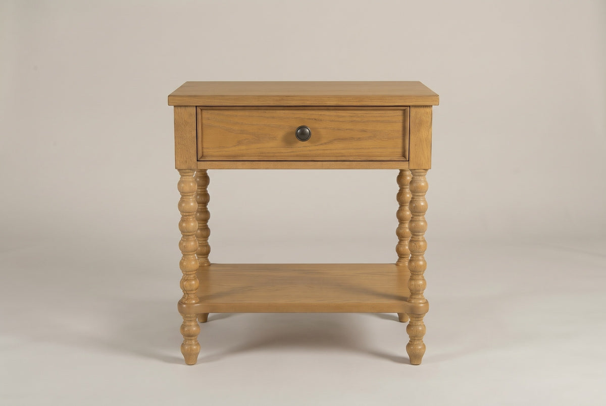 The Orgosolo Camel Oak nightstand, front view on a warm greige studio backdrop: a flat top, one drawer with one knob, four turned bobbin legs and an open shelf