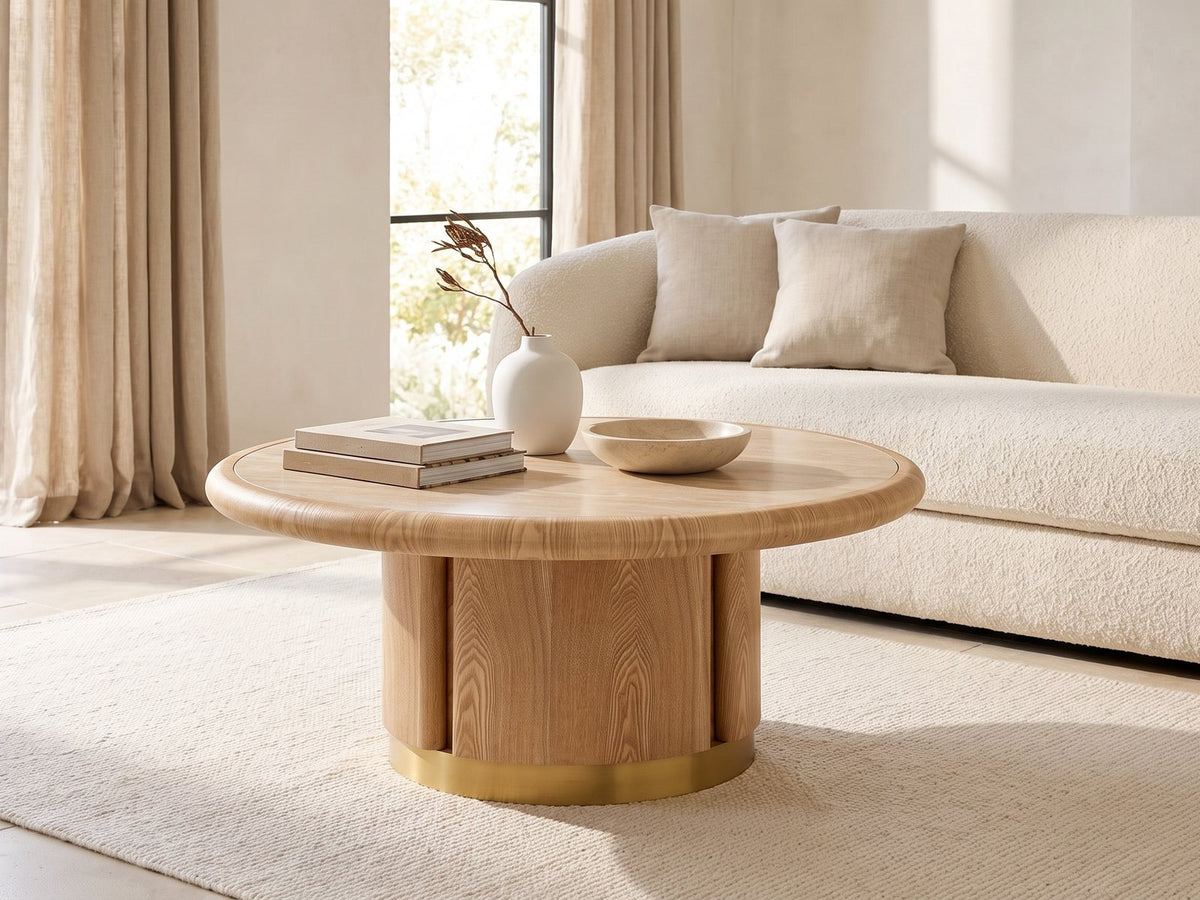 Ovada 35-Inch Round Sintered Stone and Wood Veneer Coffee Table seen from the side, styled with books, a vase and a stone bowl in front of a cream sofa in a bright plaster room