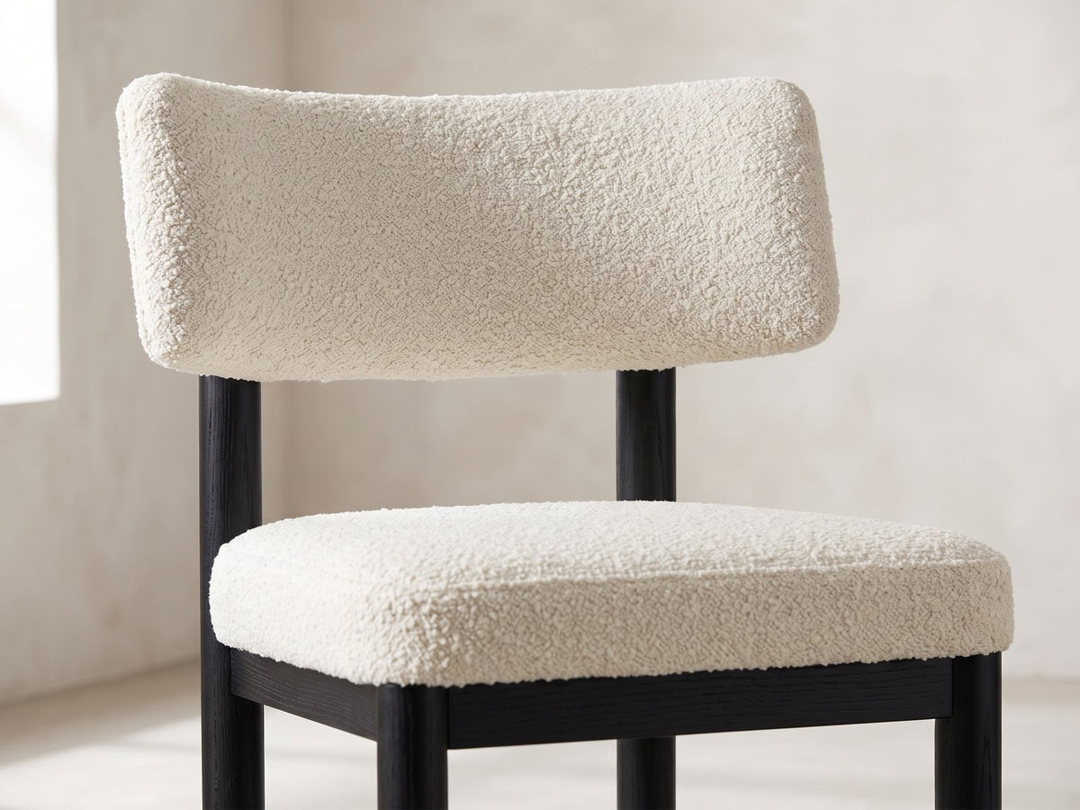 Close-up of the Palmi 70-Inch Black Oak Veneer Dining Set with 4 Boucle Chairs: the curved ivory boucle back pad and padded seat of a chair on round black legs