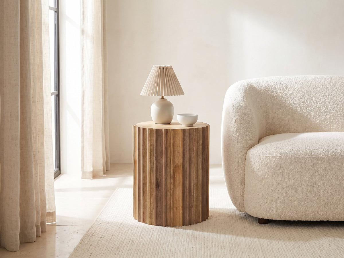 Pisticci Solid Mango Fluted Drum Side Table with a lamp and a ceramic bowl beside a cream boucle armchair in a bright plaster room