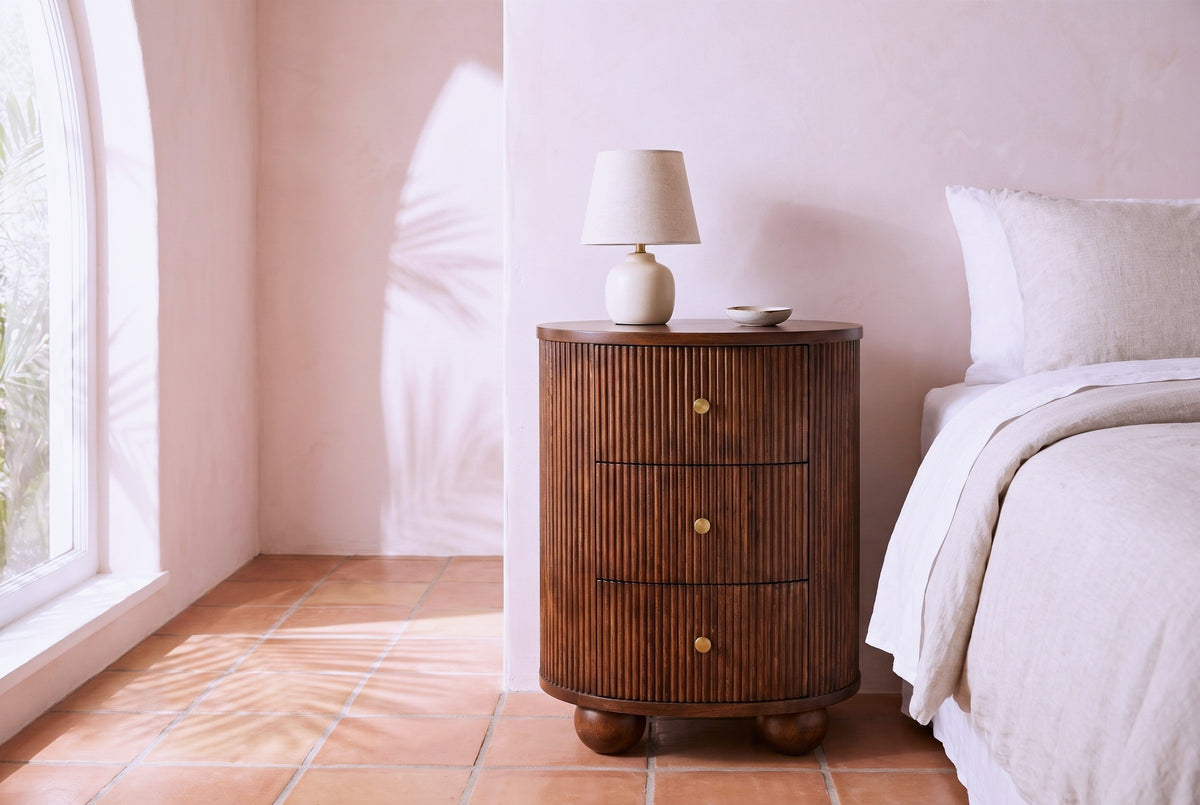 Ploaghe chestnut mango oval nightstand with a ceramic lamp beside a bed in a Los Angeles bungalow bedroom with a blush pink plaster wall and terracotta floor tiles