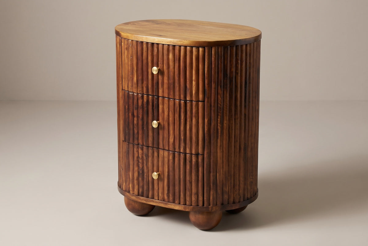 The Ploaghe oval three-drawer nightstand in chestnut mango wood with fine vertical reeds and brass knobs, three-quarter view on a light studio backdrop