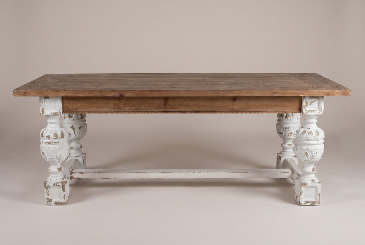 The Policoro dining table, front view of the long side on a light studio backdrop: a fir apron above four carved urn legs and one stretcher