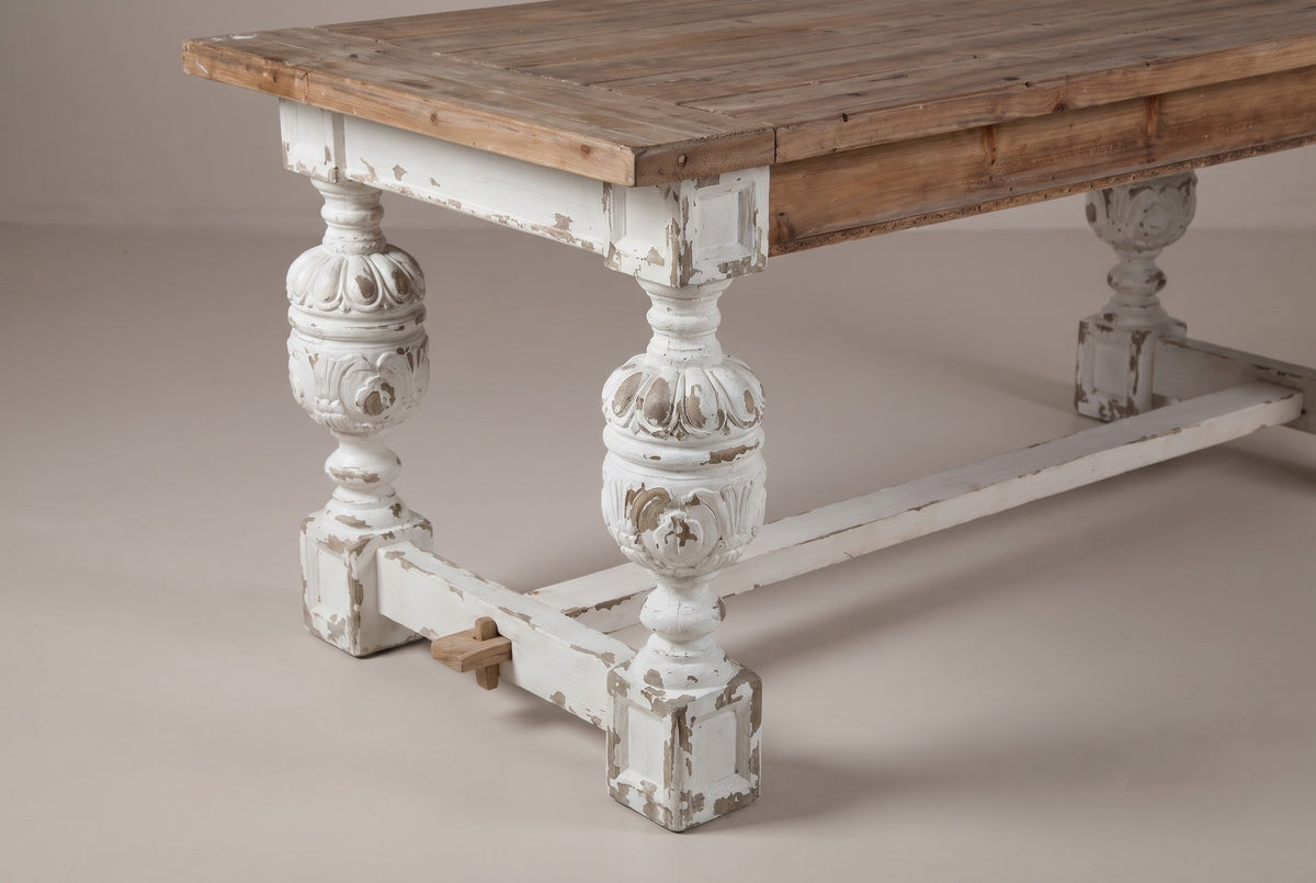 A near corner of the Policoro dining table: the fir top, a square block under it, a carved antique-white urn leg on a square block foot, the end rail with its small wooden key and the stretcher