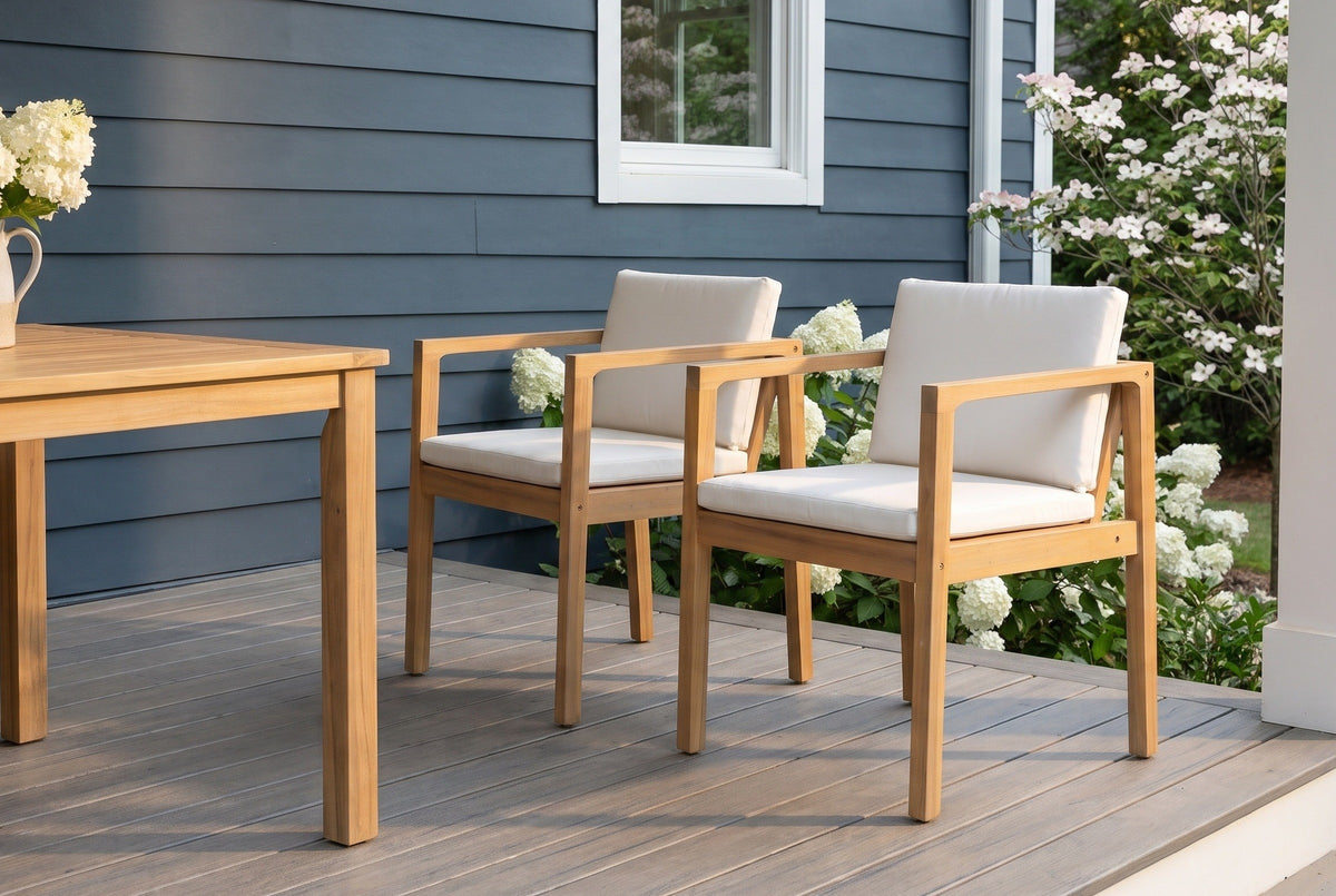 Two Pollina acacia dining armchairs on a grey porch deck beside an acacia dining table, a slate-blue shiplap wall and white hydrangeas behind (table not included)