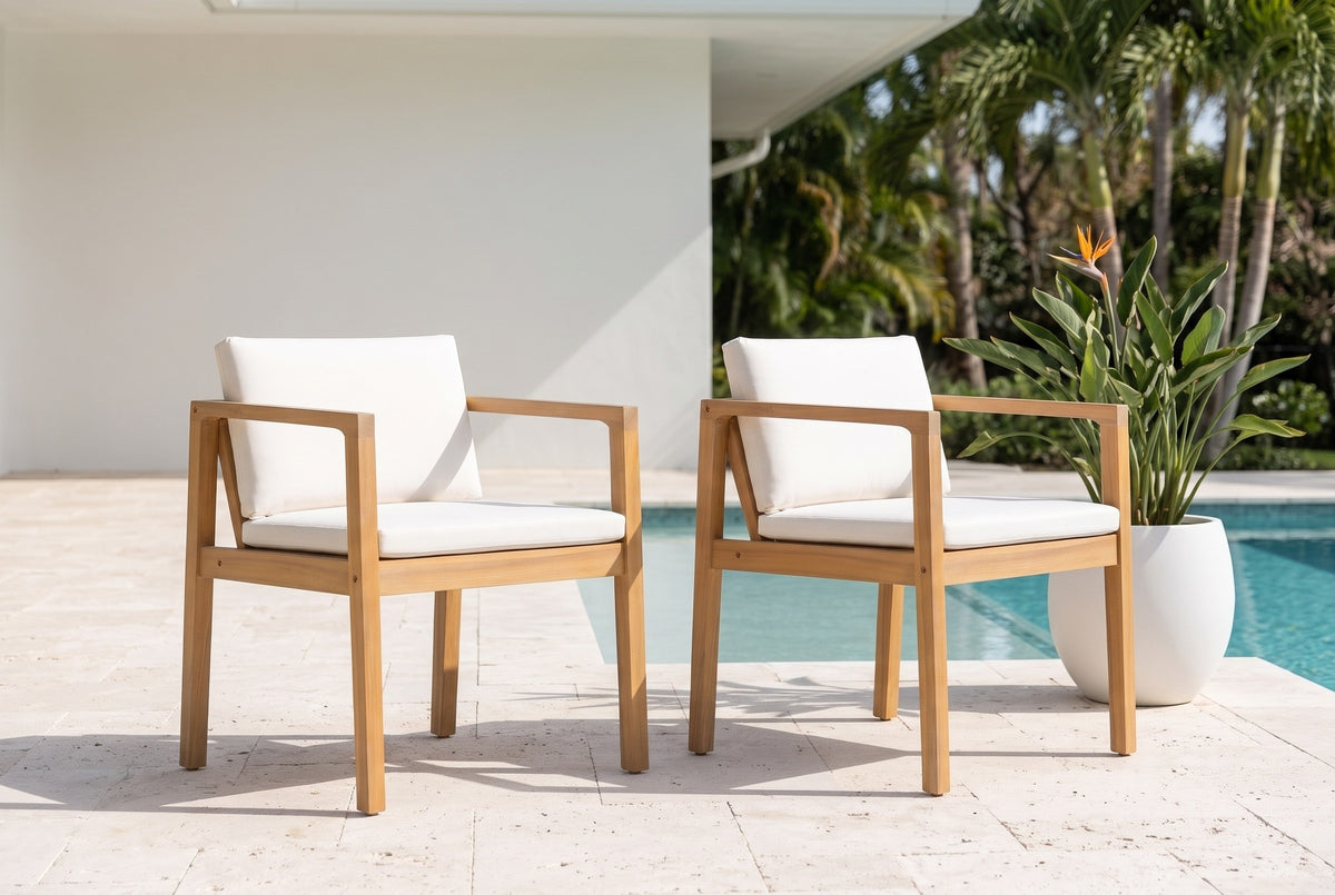 Two Pollina acacia dining armchairs on a pale coral-stone pool terrace, a white stucco wall, a pool and palms beyond (planter not included)