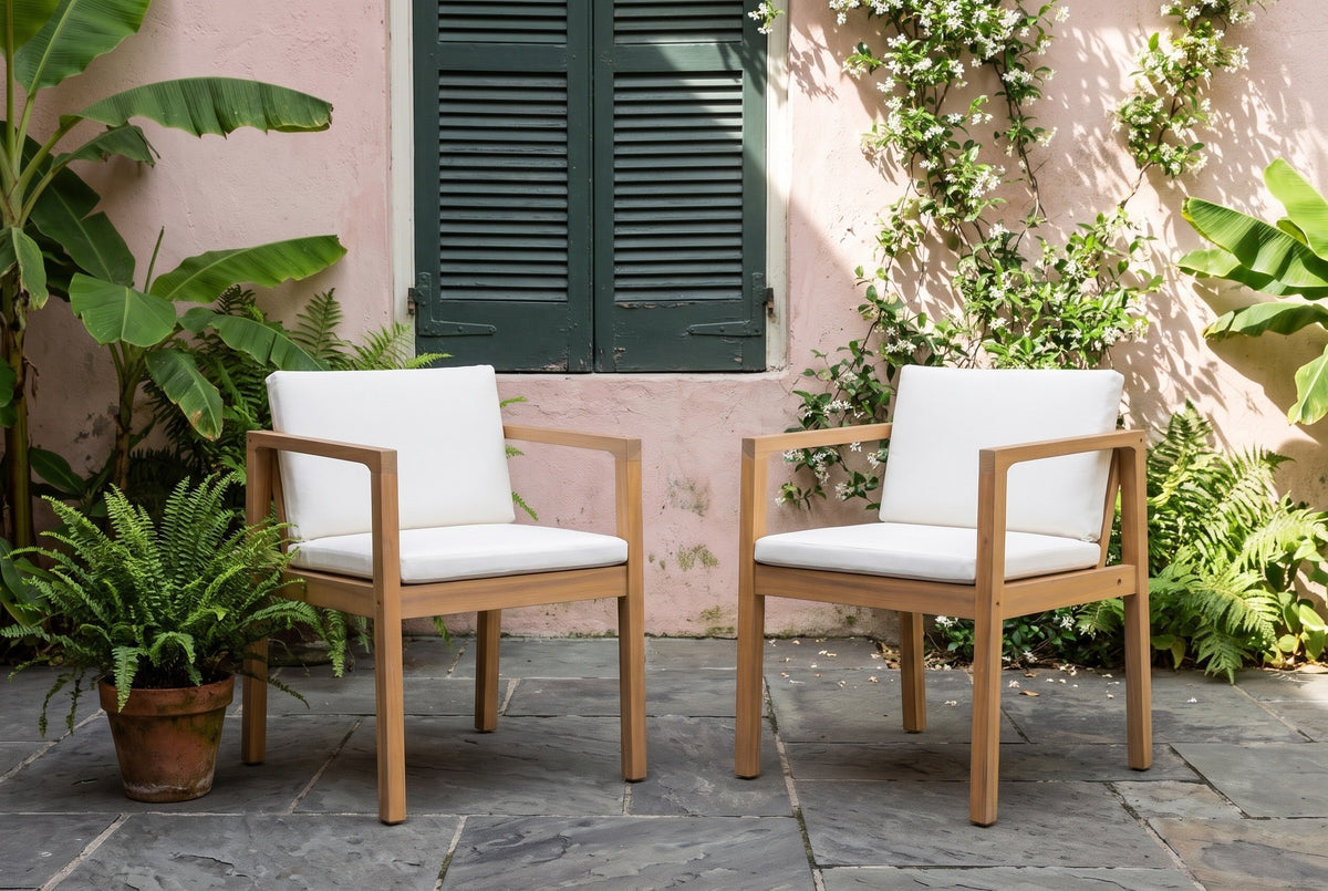 Two Pollina acacia dining armchairs on slate flagstones in a courtyard, a rose-pink plaster wall with a green shutter, ferns and banana leaves behind (planter not included)
