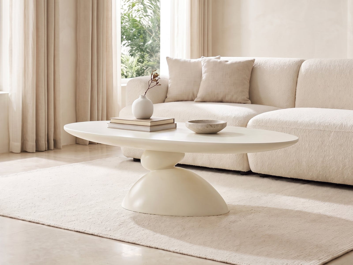 Ponza Milk White Fiberglass Pebble Coffee Table seen from the side, styled with books, a vase and a stone bowl in front of a cream sofa in a bright plaster room