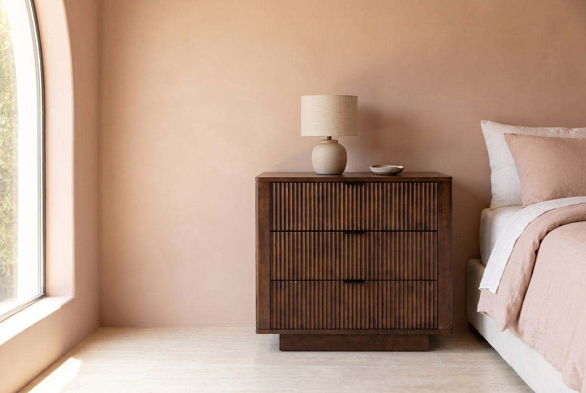 Pula reeded walnut three-drawer nightstand with a ceramic lamp beside a bed in a Los Angeles bedroom with a blush-pink lime plaster wall and an arched window