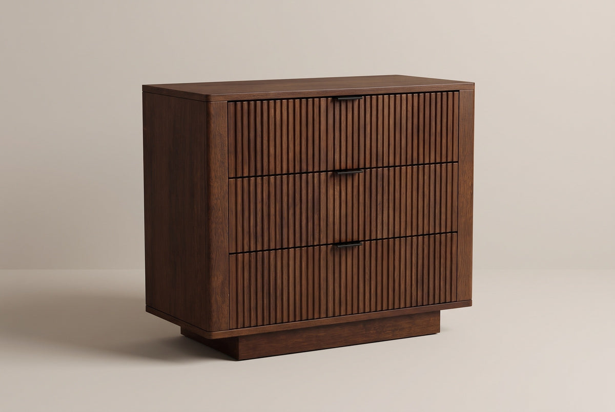 The Pula three-drawer nightstand in dark walnut with fully reeded drawer fronts and black tab pulls, three-quarter view on a light studio backdrop