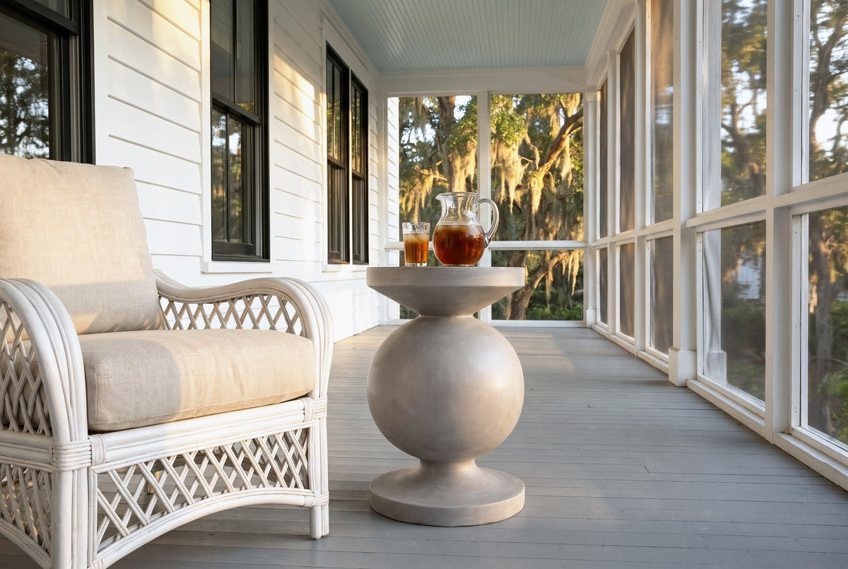 Raffadali concrete sphere pedestal side table with a pitcher of iced tea and a glass beside a white wicker armchair on a screened porch in Beaufort, South Carolina, white clapboard, grey floorboards and live oaks with Spanish moss outside (chair and decor not included)