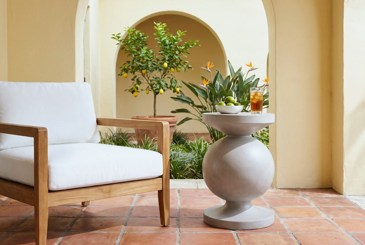 Raffadali light grey concrete sphere pedestal side table with a bowl of limes and a glass of iced tea beside a teak armchair in a Coral Gables courtyard loggia, a butter-yellow stucco arch, a lemon tree and clay tile floor (chair and decor not included)