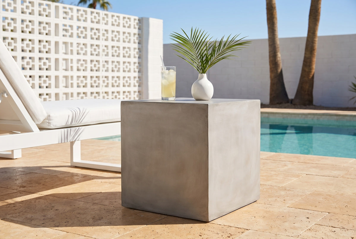 Realmonte light grey concrete cube side table with a glass of lemonade and a white vase beside a white lounge chair on a travertine pool patio in Palm Springs, a white breeze-block wall behind (chair and decor not included)
