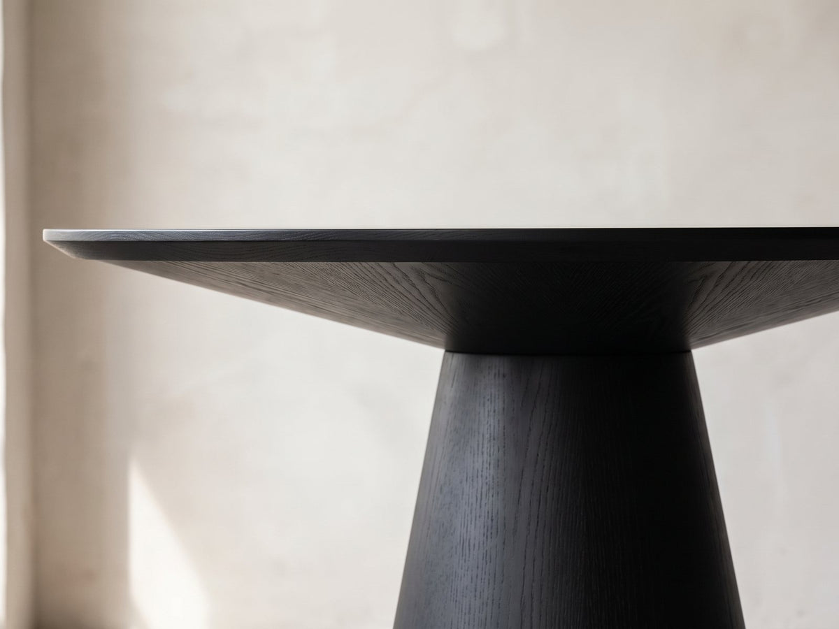 Close-up of the Savelli 47-Inch Round Black Oak Pedestal Dining Set with 4 Boucle Chairs: the slim edge and sloped underside of the round top over the black cone pedestal
