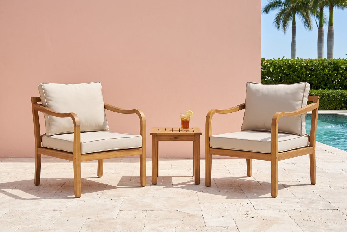 Two Savoca acacia club chairs on travertine pavers by a pool, a coral-pink stucco wall, a sea-grape hedge and royal palms behind (side table not included)