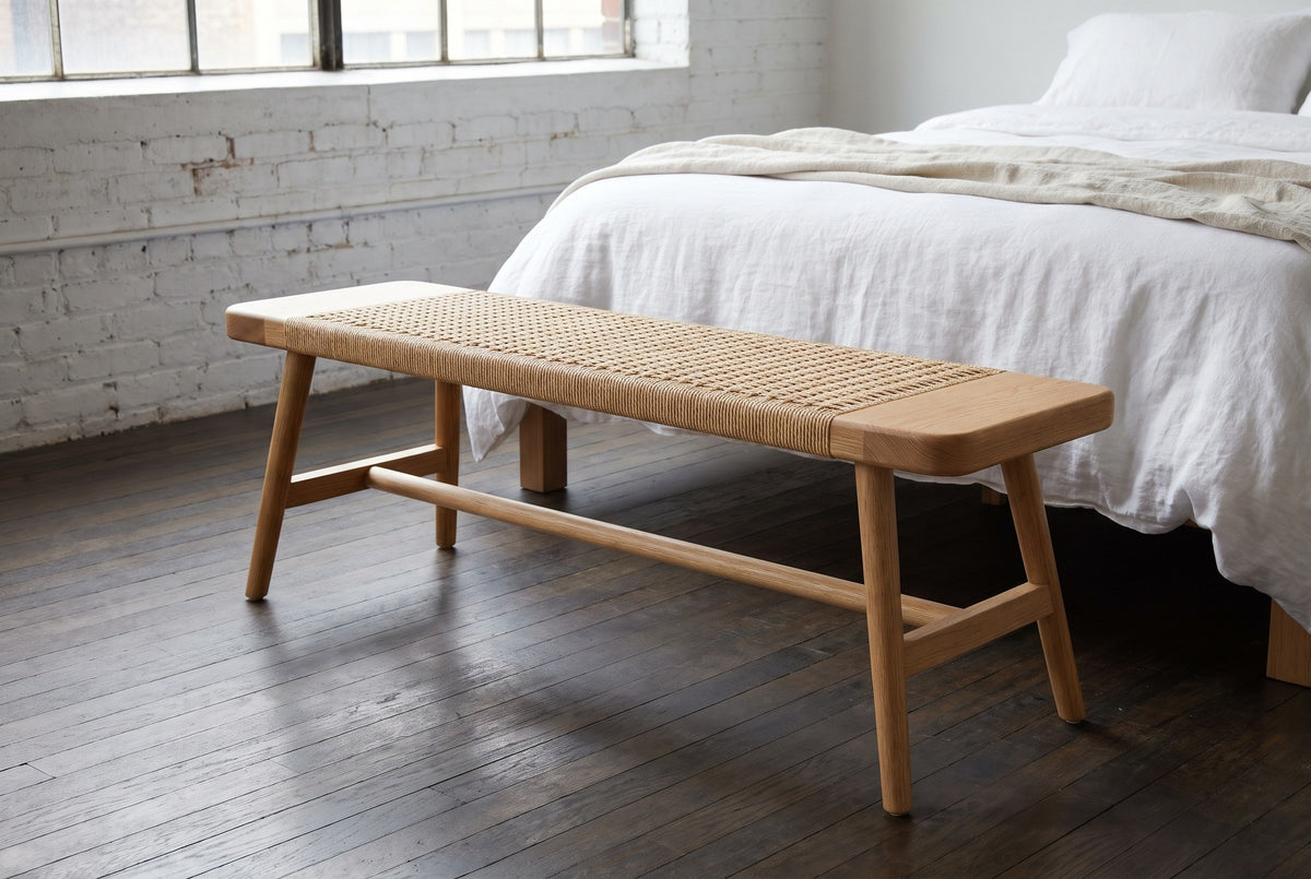 Sperlonga oak bench with a paper cord seat at the foot of a bed with white linen in a Brooklyn loft bedroom with a white-painted brick wall and dark oak floorboards (bed not included)