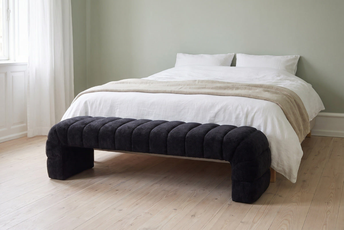 Spoleto black chenille bench at the foot of a made bed in a Copenhagen bedroom with a sage-green wall, white linen and pale oak floorboards