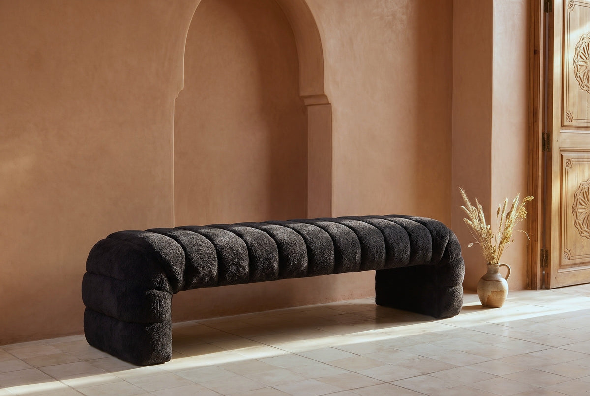 Spoleto black chenille bench against a terracotta tadelakt plaster wall with an arched niche in a Marrakech riad entry, beside a carved wooden door and a vase of dried grasses