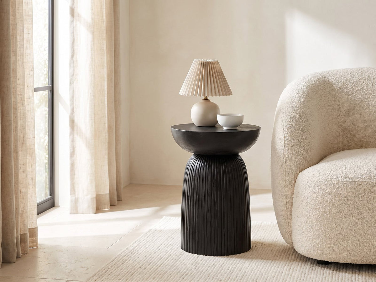 Stigliano Black Mango Bell Pedestal Side Table with a lamp and a ceramic bowl beside a cream boucle armchair in a bright plaster room