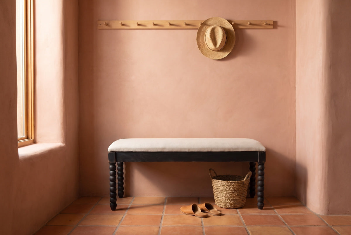 Sutri black bobbin-leg bench against a terracotta-pink adobe plaster wall in a Santa Fe entry with a saltillo tile floor, a straw hat on a peg rail, a seagrass basket and a pair of leather slippers