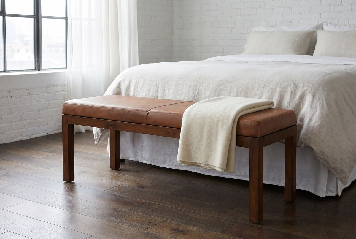 Todi antique tan leather bench at the foot of a bed with white linen bedding in a New York loft bedroom with a white-painted brick wall and a dark oak floor, a folded cream blanket on the bench