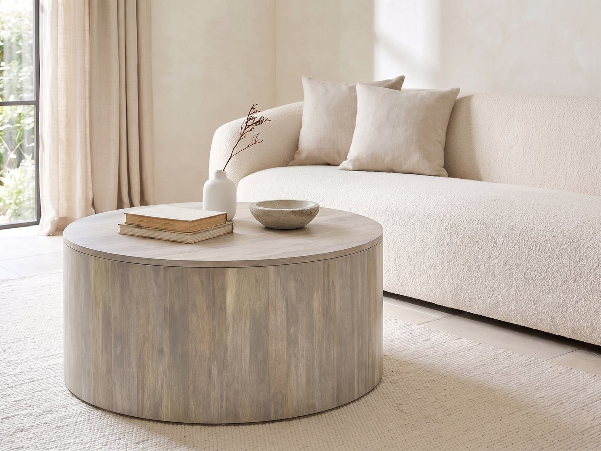 Tolentino Whitewashed Mango Drum Coffee Table seen from the side, styled with books, a vase and a stone bowl in front of a cream sofa in a bright plaster room